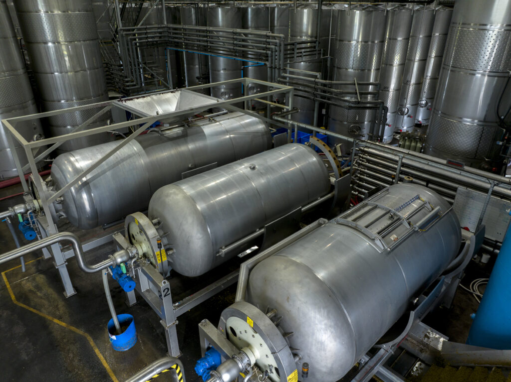 Large stainless-steel wine presses inside the winery’s production area, surrounded by tanks, pipes, and processing equipment.