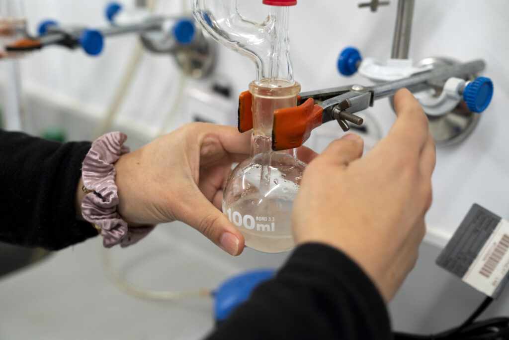Hands securing a 100ml round-bottom flask to a glass distillation setup in a winery laboratory.
